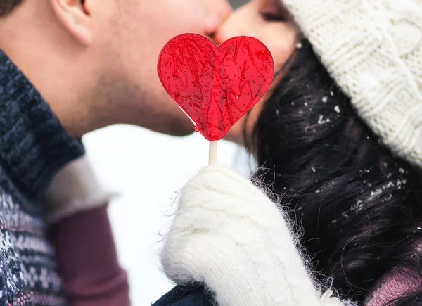 Outdoors lifestyle close up portrait of happy kissing couple in love. Holding in their hands red lollipop in shape of heart. On honeymoon. Wearing knitted hat, mittens and sweaters.  Love concept