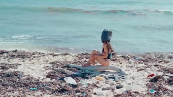 一个头上背着黑色塑料袋的女孩坐在加勒比海岸的塑料垃圾和海藻中 — 图库视频影像