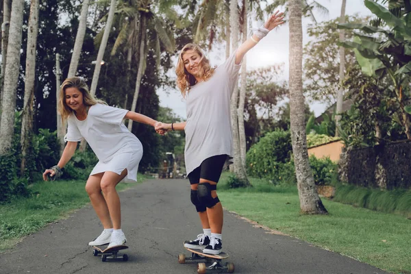 Zwei junge, schöne Frauen fahren Skateboards auf einer Straße mit Palmen — Stockfoto