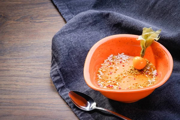 Sea buckthorn dessert in orange bowl on wooden table background with denim tablecloth. Decorated, delicious vegetarian raw dessert mousse with physalis. Restaurant menu. Copy space, close up