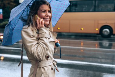 Mobil telefonda konuşurken ve bir şemsiye açık havada şehirde tutan gülümseyen genç kadın.