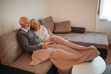 A senior couple cuddling on the living room sofa while watching television. clipart