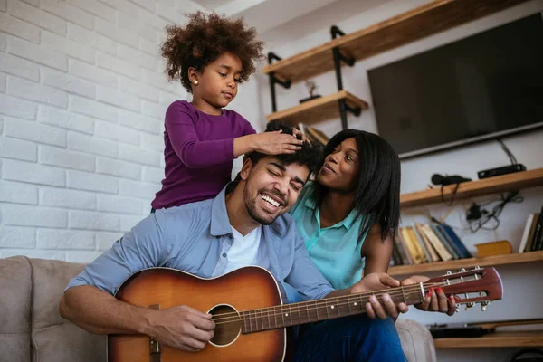 A happy black family having a good time together while the father is performing on an acoustic guitar.