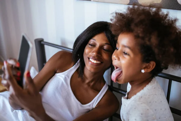 Photo of african american mother and daughter taking a picture with mobile phone in the bed.