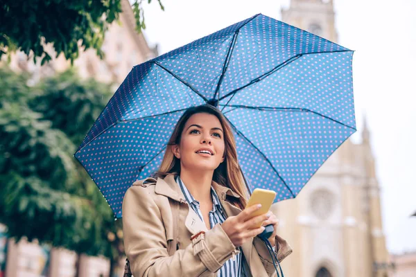 A young woman in a rain coat checking her phone while waiting for her ride under the umbrella.