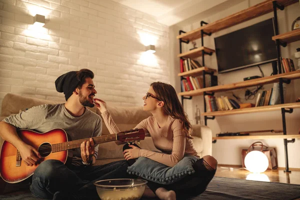 若いカップルがリラックスできる自宅で ガール フレンドは彼にいくつかのポップコーンを給餌しながら彼氏がギターを弾いています — ストック写真