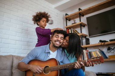 Bir gitar, karısı onu yanak ve saçlarıyla oynarken kızı öpüşme bir genç baba kadeh