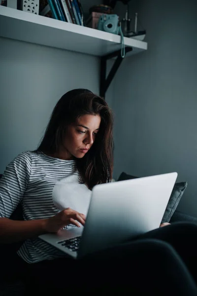 A young woman is working remotely from a dimly lit living room.