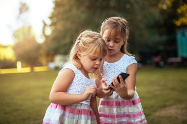 İki memnun küçük çocuklar birlikte parkta cep telefonu kullanırken vakit zevk kadeh.