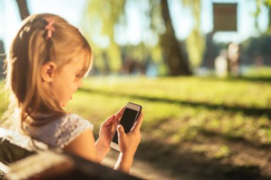 Sevimli küçük kız parkta bir bankta otururken bir cep telefonu kullanarak.