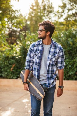 Yakışıklı adam şehirde yürüyüş güneş gözlüklü bir longboard taşıyan.