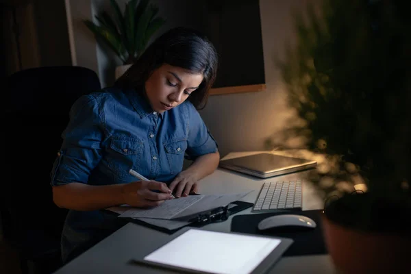 Photo of a young woman sitting at the computer table and working late at night.