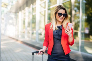 Bir bavul çekerek ve açık havada cep telefonuyla gülümseyen bir iş kadın portresi.