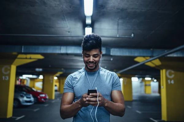 Portrait of african american athlete man listening to music and using mobile phone while working out in the underground parking.