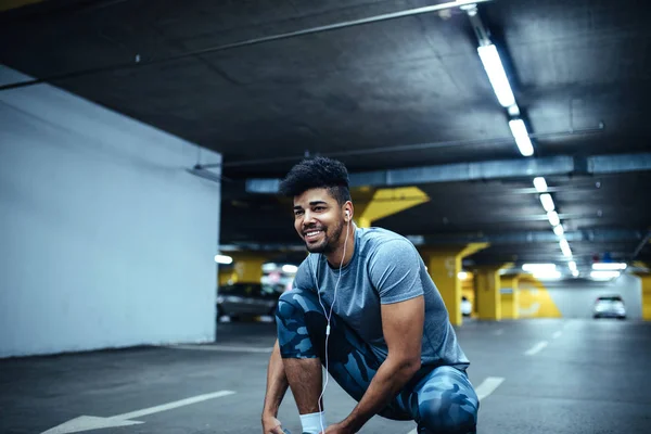 Portrait of happy african american athlete man tying shoes before run in the underground car parking while listening to the music.