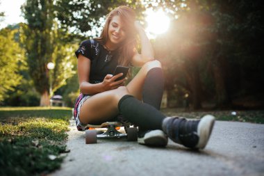 Cep telefonu kullaranarak ve gün batımında açık longboard üzerinde oturan neşeli genç kadın.