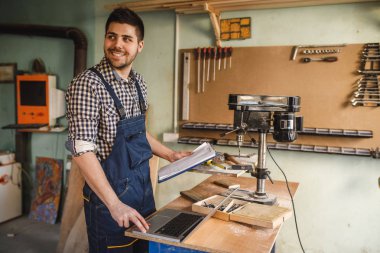 Gülümseyen genç marangoz atölyesinde dizüstü kullanarak portresi.