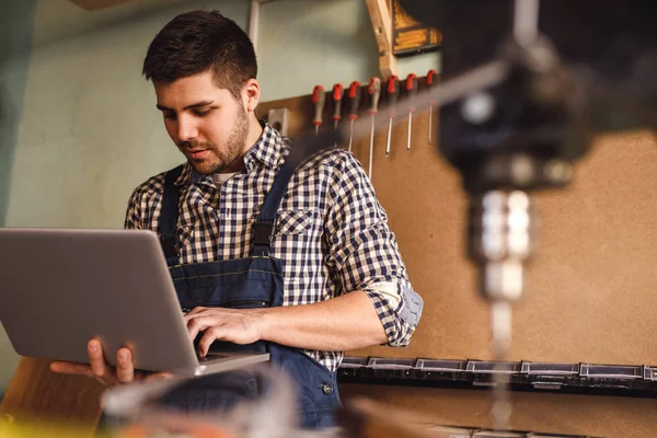 Yakışıklı bir marangoz atölyesinde çalışırken dizüstü kullanarak atış.