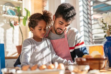 Baba ve kızı masada oturan ve pişirmeye hazırlanıyor. Önlük giyen ve babadır.