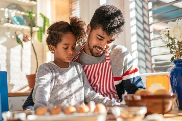 Baba ve kızı masada oturan ve pişirmeye hazırlanıyor. Önlük giyen ve babadır.