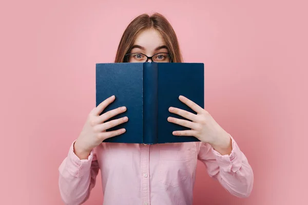 Ragazza emotiva in occhiali nascondendo faccia dietro libro aperto — Foto Stock