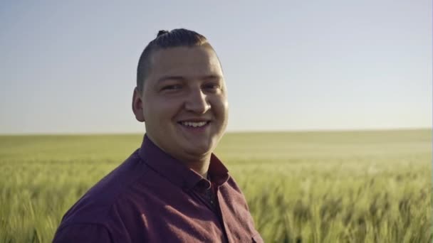 Крупный план портрета улыбающегося человека, стоящего на зеленом пшеничном поле на закате и смотрящего в камеру - замедленная съемка — стоковое видео