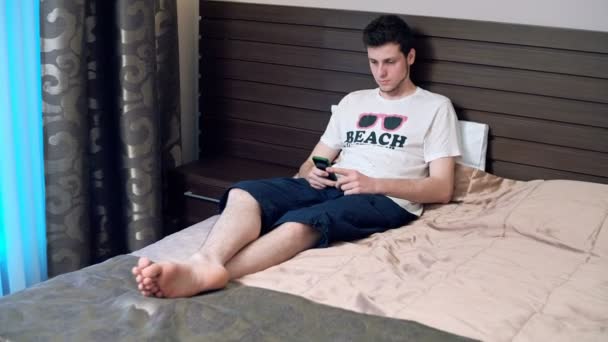 Sad Young Man with Cellphone sitting on the bed. RAW video record, — Stock Video