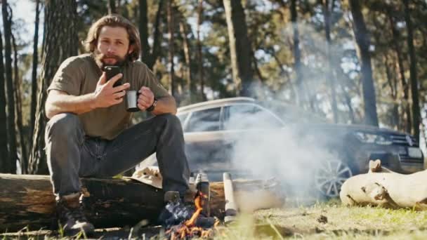 Joven hombre maduro hermoso sentado en el fuego en el bosque de pinos de verano y beber té. El coche está de pie junto a él . — Vídeos de Stock