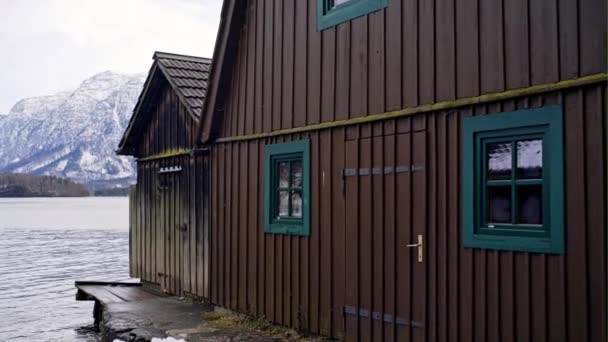 Eenvoudige houten plank gebouw geplaatst aan oever van niet bevroren meer met hoge besneeuwde bergen op de achtergrond in de winter in Hallstatt, Oostenrijk — Stockvideo