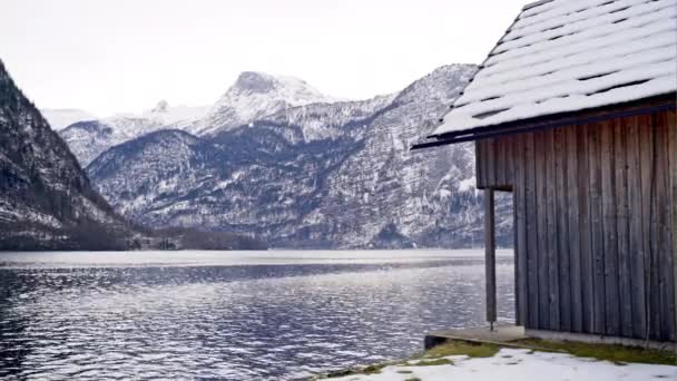 Ancien bâtiment en planches de bois placé au bord du lac avec de magnifiques hautes montagnes avec de la neige en arrière-plan en hiver à Hallstatt, Autriche — Video