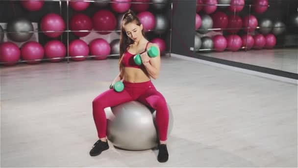 Mujer joven entrenando con pesas mientras está sentada en la pelota de ejercicio en el gimnasio . — Vídeos de Stock