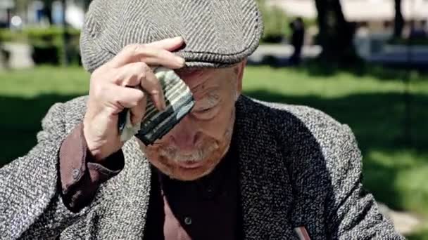 Горячий усталый старик с тростью в парке вытирает пот со лба платком, а потом снимает кепку. — стоковое видео