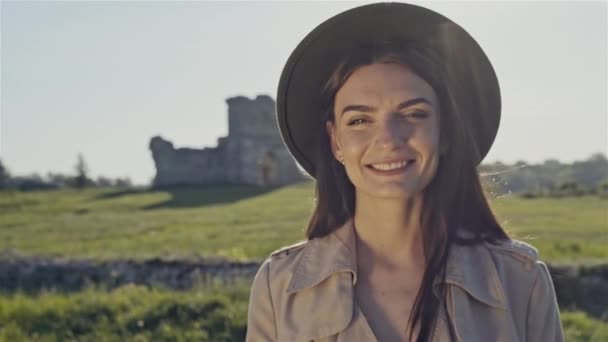 Mooie brunette in hoed en vacht permanent in het zonnetje op de achtergrond van de landelijke natuur achtergrond. — Stockvideo