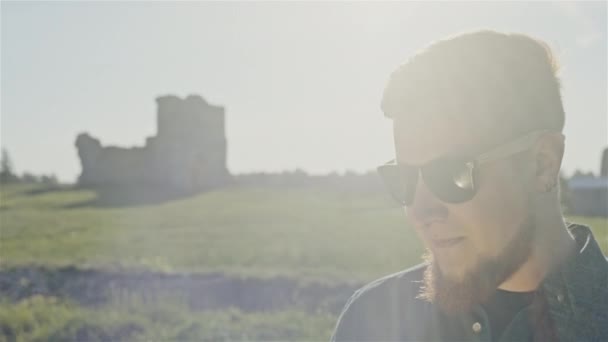Young bearded trendy man in sunglasses standing in sunlight on background of green terrain in countryside — Stock Video