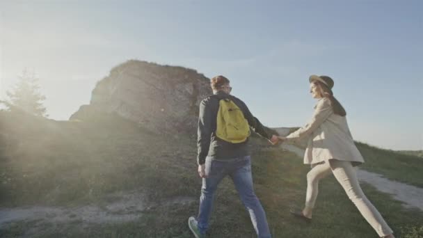 Vista trasera de la feliz pareja elegante cogida de la mano y caminando por el terreno rural con roca bajo el sol brillante — Vídeo de stock