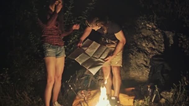 Un par de jóvenes excursionistas leyendo un mapa a la luz de una hoguera ardiendo por la noche inclinándose hacia el resplandor de las llamas . — Vídeo de stock