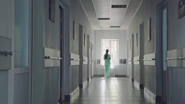 在医院通道末端的手术磨砂的妇女站在窗口前面的后退视图 — 图库视频影像