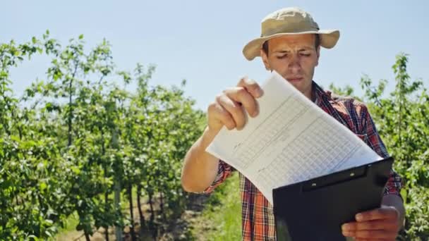 Çiftçi bir meyve bahçesinde kameraya gülümseyen kolunun altında Pano'ya koymadan önce bir elektronik tablo denetimi genç meyve ağaçları ile ayakta. — Stok video