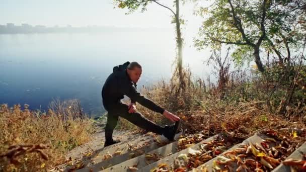 Joven en ropa deportiva haciendo ejercicio de calentamiento mientras está de pie en las escaleras cerca de lago tranquilo en el día soleado de otoño — Vídeos de Stock