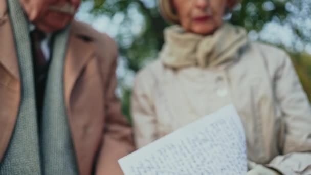 Senior Mann und Frau in eleganten Outfits lächeln und lesen alte Briefe zusammen, während sie auf einer Bank im herrlichen Herbstpark sitzen. — Stockvideo