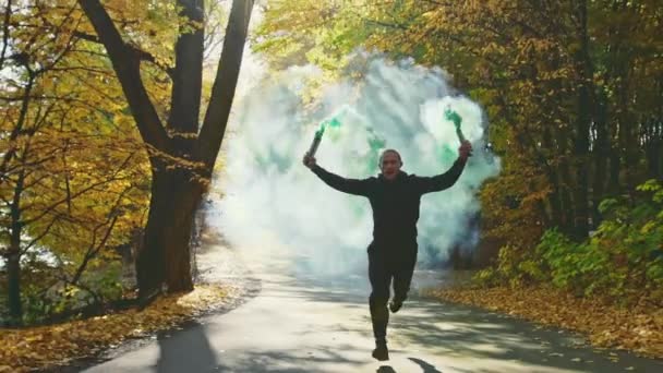 Adatto uomo atletico che corre lungo una strada forestale con fogliame colorato autunno tenendo in alto ardenti razzi fumo finale — Video Stock