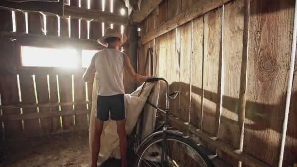 Adolescente campesino en sombrero de paja quitando la cubierta de la bicicleta vintage y rodando fuera del hangar de madera - cámara lenta — Vídeos de Stock
