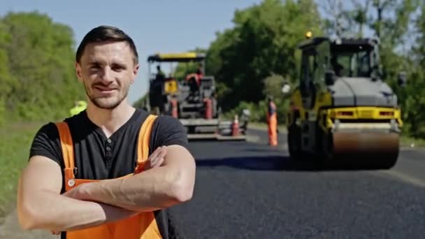 Portrait d'un travailleur heureux qui répare les routes sur le fond du rouleau d'asphalte et d'autres équipements. Un employé heureux regarde la caméra . — Video
