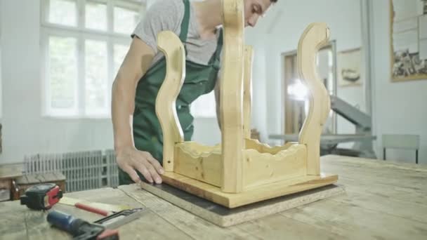 Ung man i övergripande arbetar i snickeri butik står vid bordet och skapa trästol. — Stockvideo