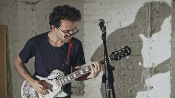 Homme décontracté dans des lunettes debout contre un mur de briques blanches et jouant de la guitare sous les projecteurs — Video