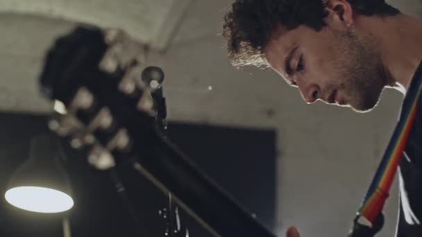 Side view of young modern casual man playing guitar while standing in studio with microphone in back lit of lamp — Stock Video