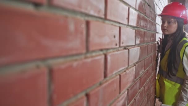 Ragazza in casco edificio protettivo e gilet controlla la qualità del lavoro sulla costruzione di un muro di mattoni. I bambini svolgono una professione da adulti . — Video Stock