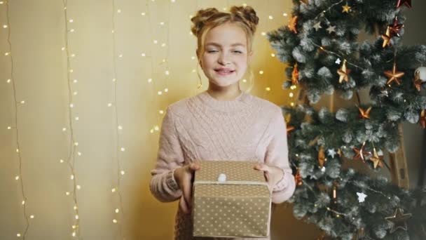 Piccola ragazza allegra che dà scatola regalo mani tese a macchina fotografica in piedi in luci incandescenti al chiuso — Video Stock