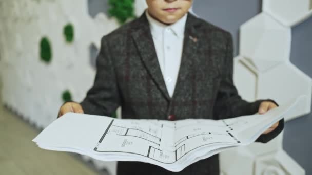 Ein komischer kleiner Junge, der sich als Projektleiter verkleidet, schaut sich in einem Büro-Setting die Pläne an, bevor er seine Zustimmung gibt — Stockvideo