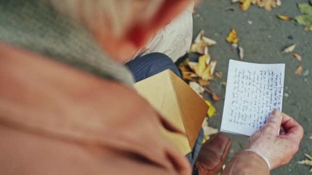 Uma vista sobre o ombro de uma pessoa idosa abrindo uma carta manuscrita em um parque com folhas de outono — Vídeo de Stock